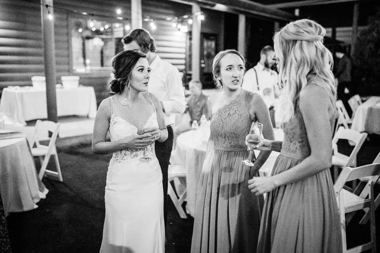 Bride and two bridesmaids talking at a casual outdoor wedding reception with string lights and a log cabin.