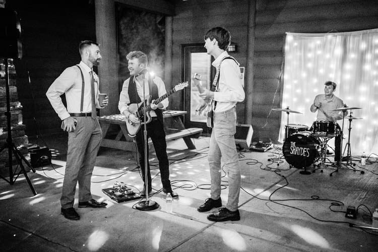 Four-man band performs at a wedding reception. Two guitarists, a drummer, and a guest stand in a rustic venue. String...