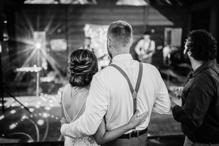 Couple from behind, embracing, watching a band perform on stage with string lights and floor lighting. Black and white...