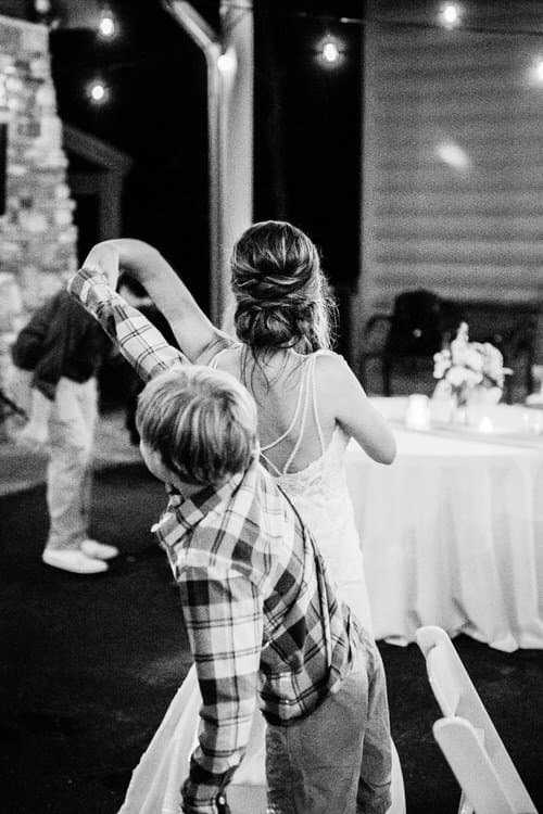 Bride in wedding dress with child in plaid shirt dancing outdoors at night, string lights overhead. Moose Hollow Lodge venue.