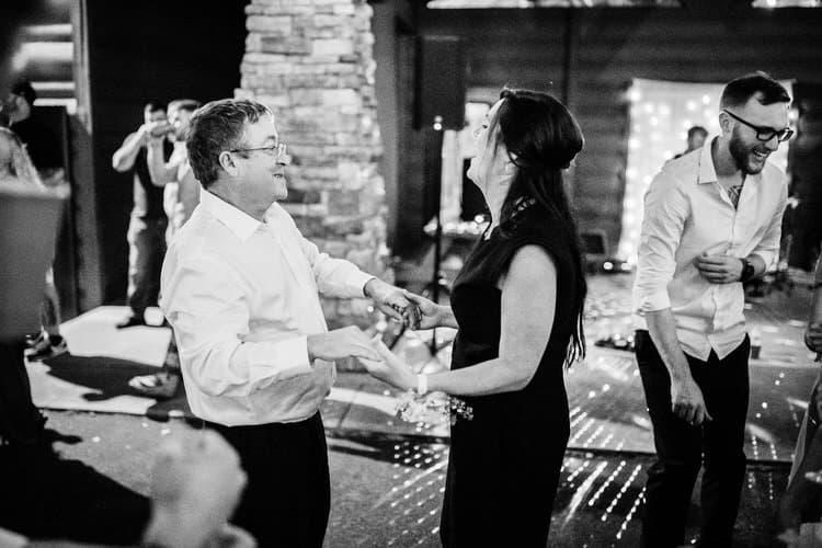 Guests dancing at a wedding reception. A man in glasses and a woman in a dress hold hands on a lit dance floor. Black and...